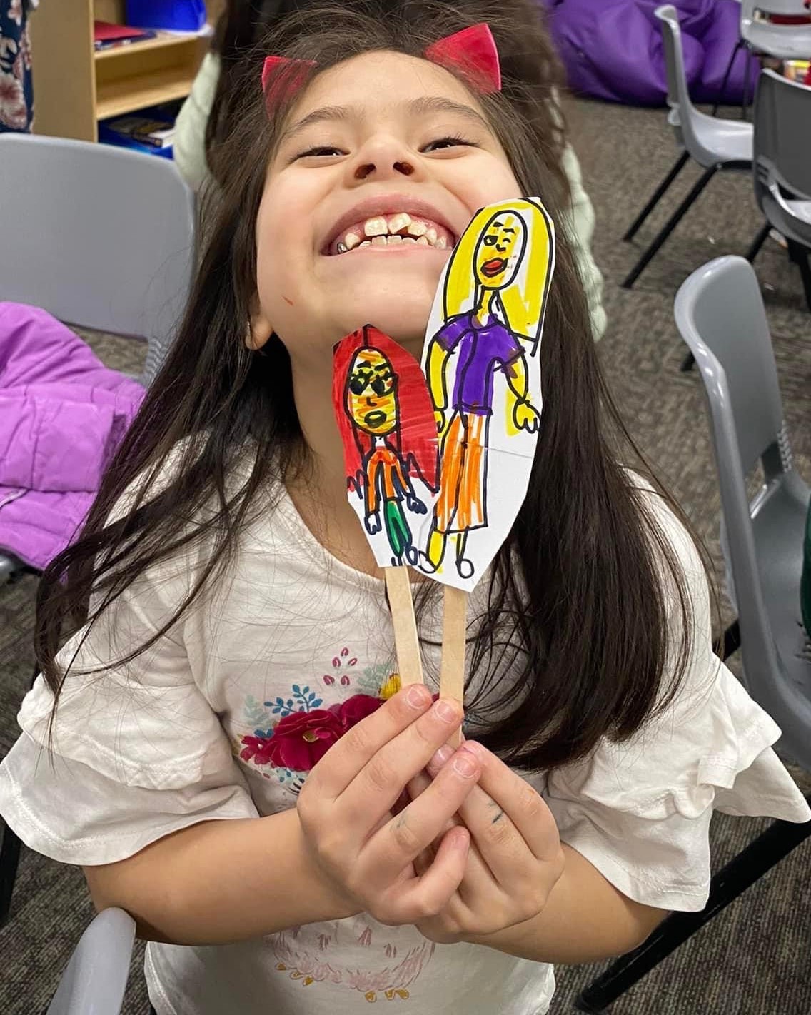 A grinning child holds up two hand-drawn paper puppets on craft sticks she made during a Heard workshop.