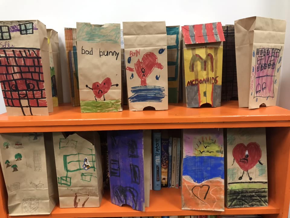 Hand-decorated paper bags shaped like houses and storefronts line an orange bookshelf after a youth art workshop.