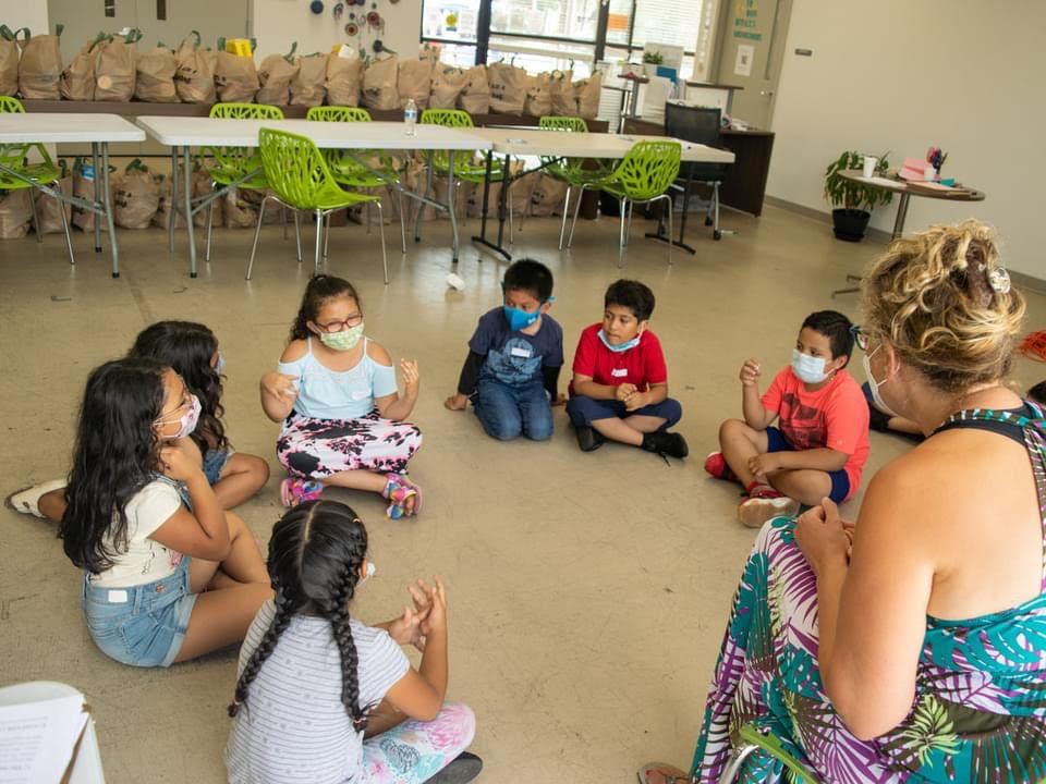 A group of children sits in a circle on the floor with a teaching artist leading an improv game.