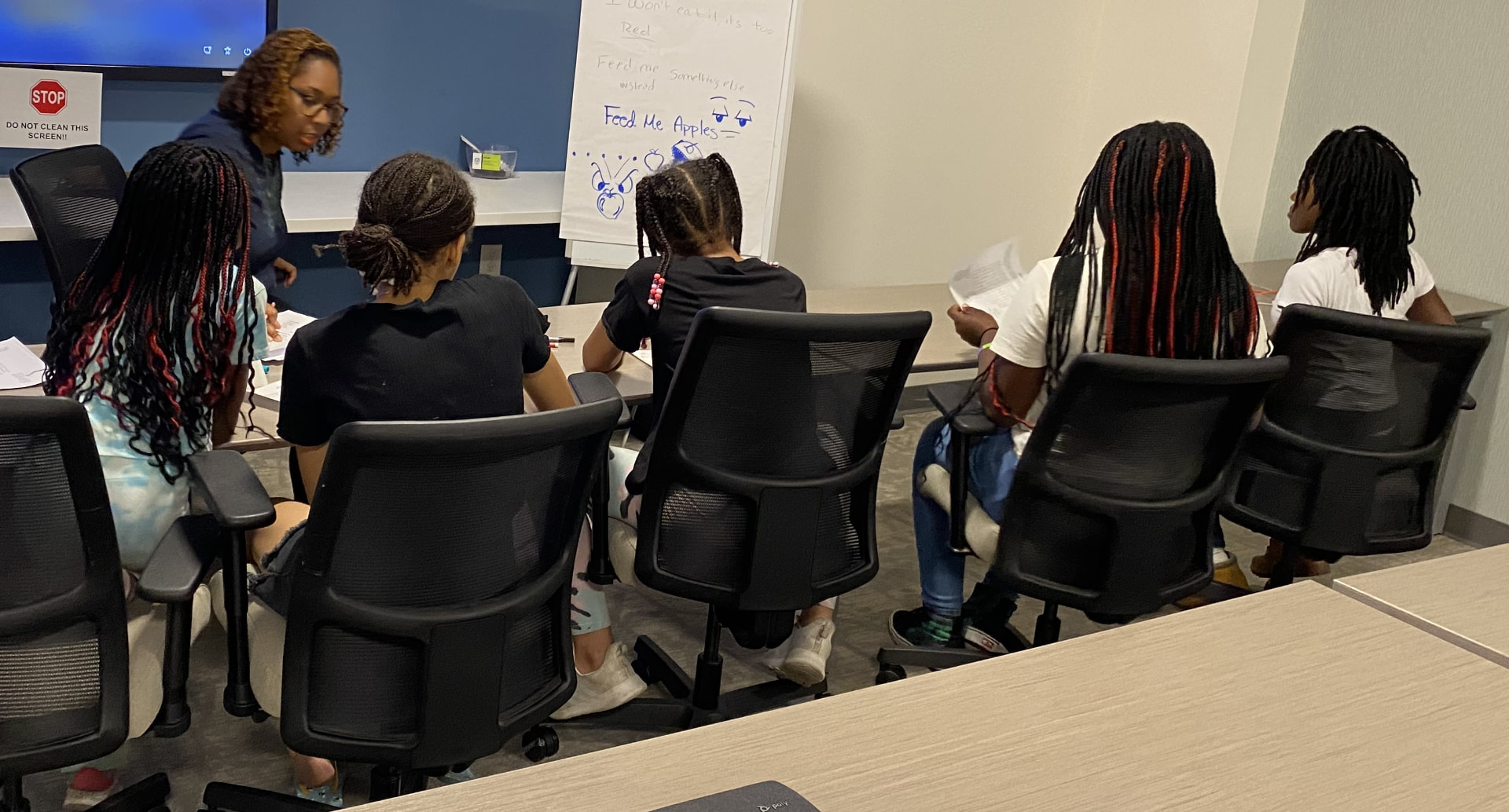 Students write at a table beside a whiteboard during a superhero-themed storytelling workshop.