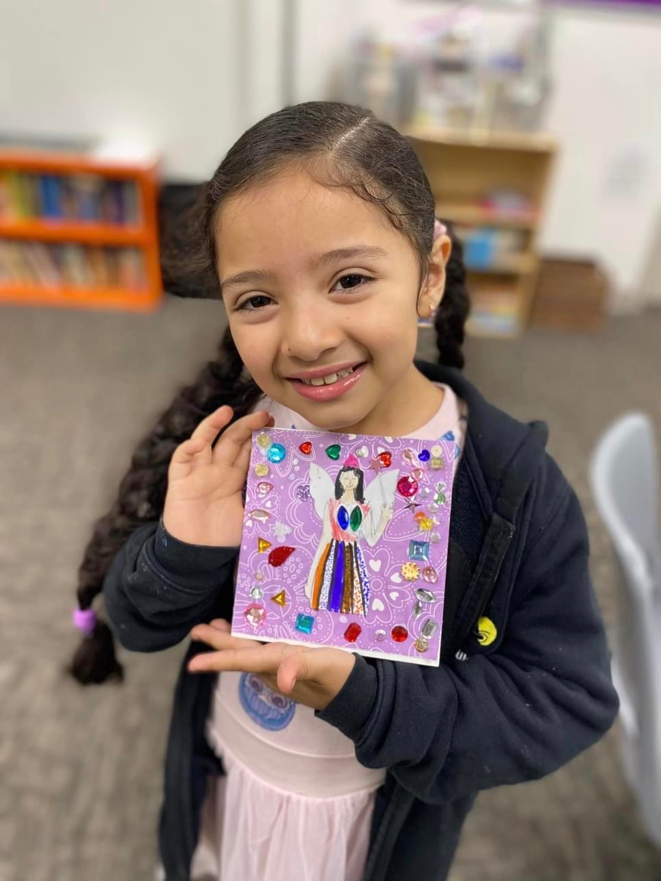 A child proudly displays a purple, jewel-covered handmade card she created in a Heard art session.