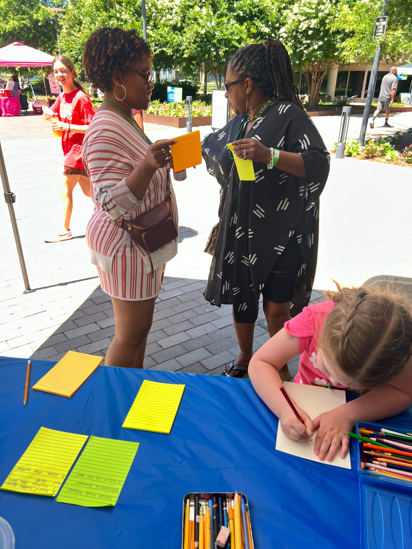 Two people talk at an outdoor community festival beside a Heard writing table stocked with paper and pencils.