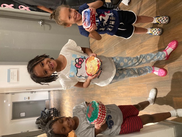 Three young children stand in a hallway smiling and holding painted boxes they decorated in a Heard art class.